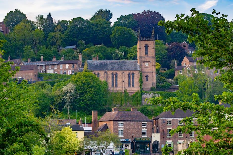 A scenic view of a small village with a prominent stone church featuring a tall clock tower nestled on a green hillside. Surrounding the church are residential houses with brick facades, nestled among