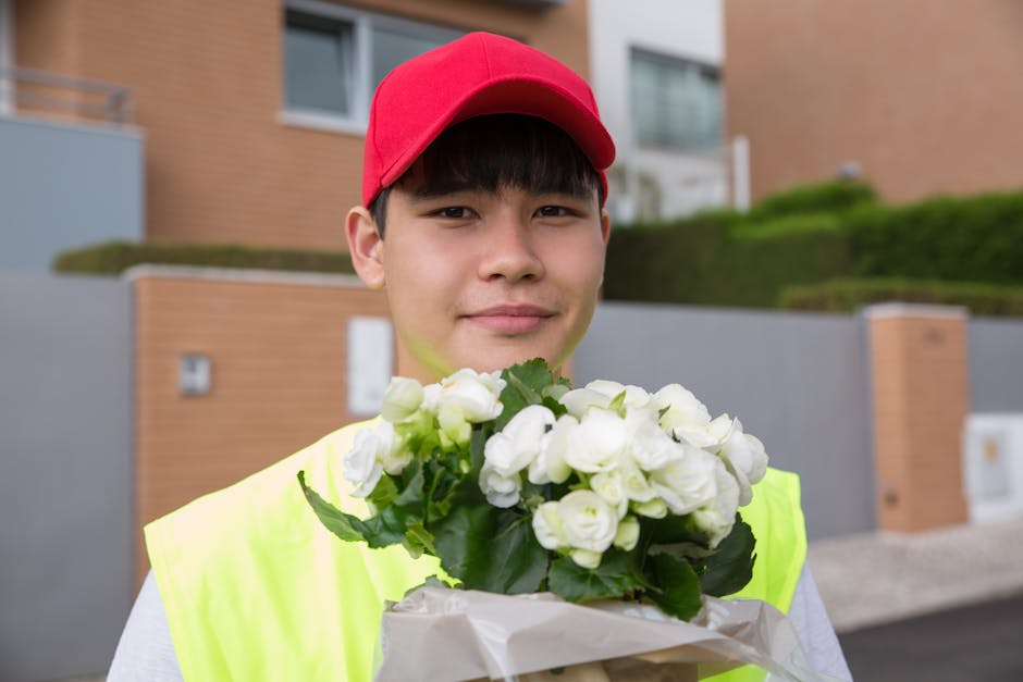 A young male flower delivery courier wearing a red cap and high-visibility yellow vest holds a bouquet of fresh white roses with green leaves, wrapped in white paper. He is standing outdoors in front 