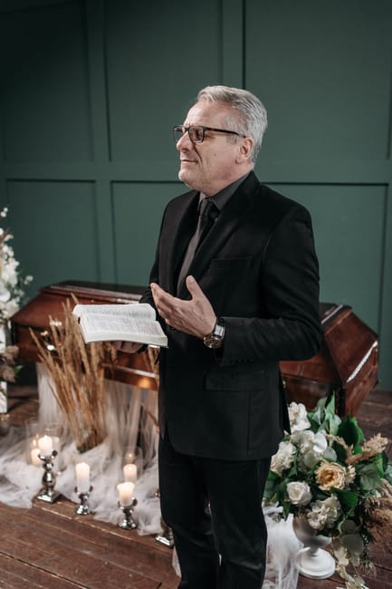 A mature man with gray hair and glasses, dressed in a black suit with a black shirt, stands in a solemn setting holding an open book, likely at a memorial or funeral service. Behind him, there's a gre