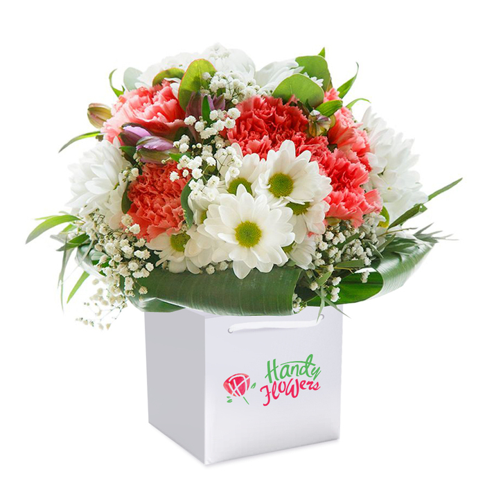 Arranged with an eye for local elegance, this vibrant bouquet from a skilled St Luke's florist stands tall within a sleek white square gift bag, the kind that hints at celebration and thoughtful giving. At the heart of the bouquet lie coral and peach carnations, recognized by their textured, ruffled petals that seem to ripple in the soft, even lighting-reminding one of the inviting flower stalls along Central Street. Intermingled are crisp white daisies with bright lime-green centers that evoke the lively palettes of local gardens, while delicate clusters of baby's breath introduce an airy, almost cloud-like contrast. Strategic whispers of purple-tinged unopened buds are tucked artfully on the left, giving depth and a sense of anticipation to the composition. Surrounding this medley is a rich collar of broad, deep-green foliage, their glossy finish reflecting the mature trees dotting St Luke's Churchyard. The bouquet's expertly rounded shape, designed for visual impact, exudes freshness and generosity, ready to lift the spirit of any recipient-be it for a birthday, an anniversary, or to send sincere cheer to a flat near the Barbican. The gift bag's soft white ribbon handles promise both style and convenience, echoing the thoughtful attention to detail of the local St Luke's floral community. Captured against a bright, minimalist background, the arrangement glows with a sense of new beginnings and heartfelt connection, a testament to the artistry and care found in every St Luke's flower delivery.